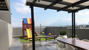 Una terraza en la azotea con zona de juegos, que incluye tobogán y columpios, y un área de comedor con sillas y una mesa.