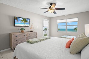A bedroom with a bed, a dresser, a television, a ceiling fan, a window with a view of the ocean, and a door with a view of the outdoors.