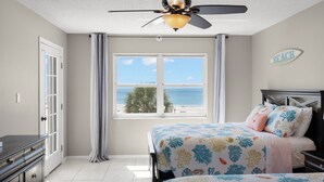 Une chambre à coucher comprenant un lit, une table de chevet, une commode, un ventilateur de plafond, une fenêtre donnant sur la plage et un panneau mural sur lequel est écrit le mot « PLAGE ».