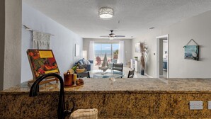 Une cuisine dotée de plans de travail en granit, d’une serviette en macramé suspendue, d’un ventilateur de plafond et offrant une vue sur le salon, où l’on aperçoit une maquette de voilier.