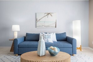 A room with a blue sofa, a white vase, a bowl with seashells, a beach-themed painting, and a lamp.