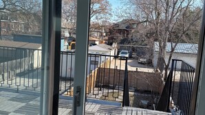 Vista de um quarto com porta de vidro deslizante para um deck externo, com piso de madeira e guarda-corpo de metal.