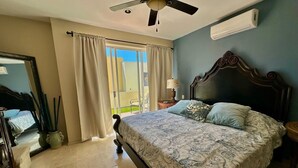 A bedroom with a bed, a ceiling fan, an air conditioning unit, a window with curtains, and a view of the outdoors.