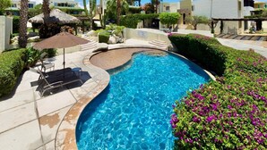 A swimming pool with a curved edge, surrounded by palm trees, a thatched-roof structure, and a flower bed.
