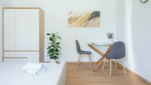 A room with a bed, a desk with two chairs, a plant, and a painting on the wall.