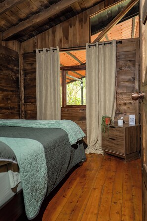 Une pièce de cabane en bois comprenant un lit, des rideaux, une fenêtre, un petit bureau avec une plante et une porte.