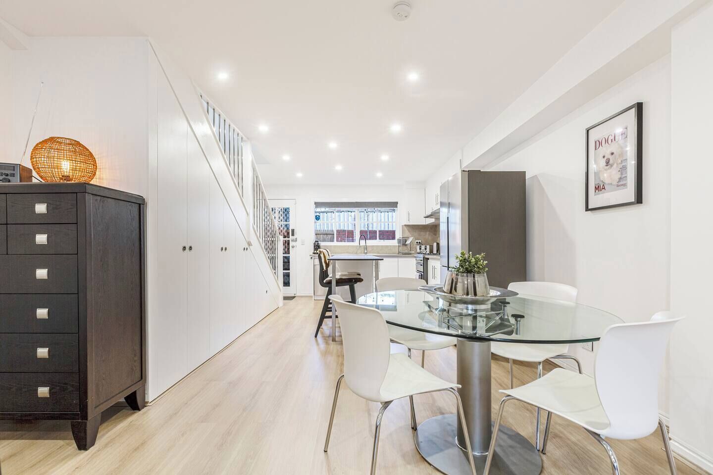 A modern kitchen with a glass dining table, white chairs, and a dark wooden cabinet.
