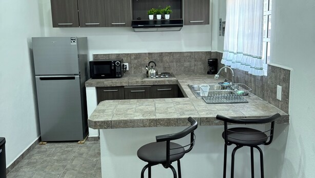 A compact kitchen with a refrigerator, microwave, kettle, and bar stools.