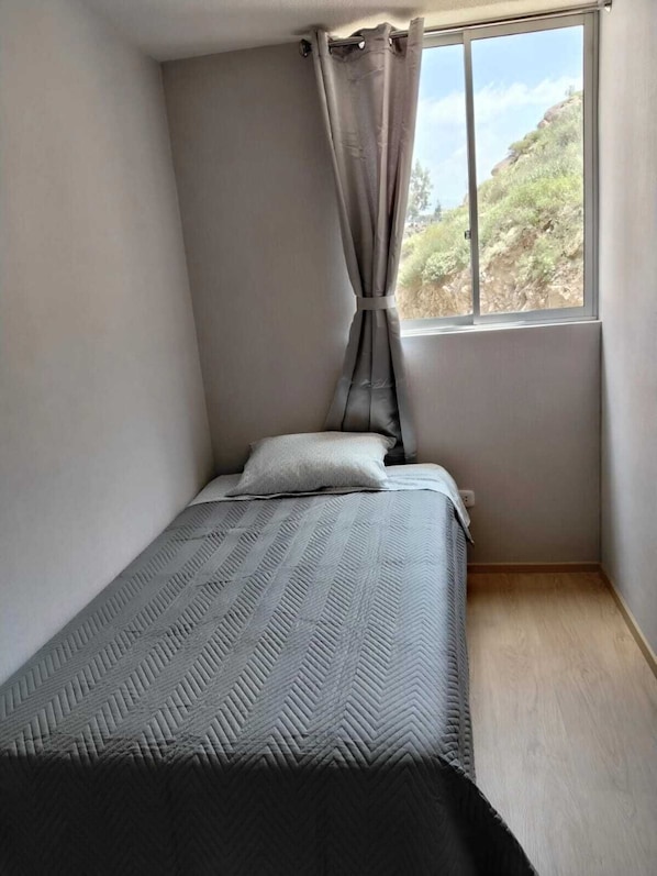 A small, single-bed room with a window, a curtain, and a plain wall.