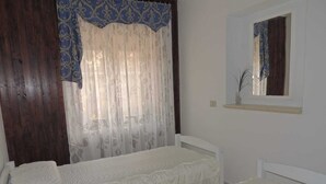 A bedroom with two beds, a window with sheer curtains, a mirror above the bed, and a wooden wall. - Vacation apartment in Santa Maria Navarrese, beautiful view (Santa Maria Navarrese)