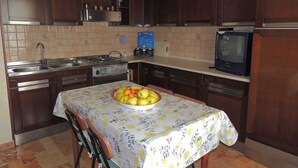 A kitchen with dark wooden cabinets, a tiled backsplash, a stove, a sink, a dining table with a fruit bowl, and a television. - Vacation apartment in Santa Maria Navarrese, beautiful view (Santa Maria Navarrese)