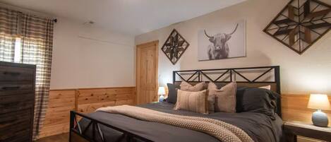 A bedroom with a bed, wooden headboard, a nightstand, a lamp, a window with curtains, and wall art.