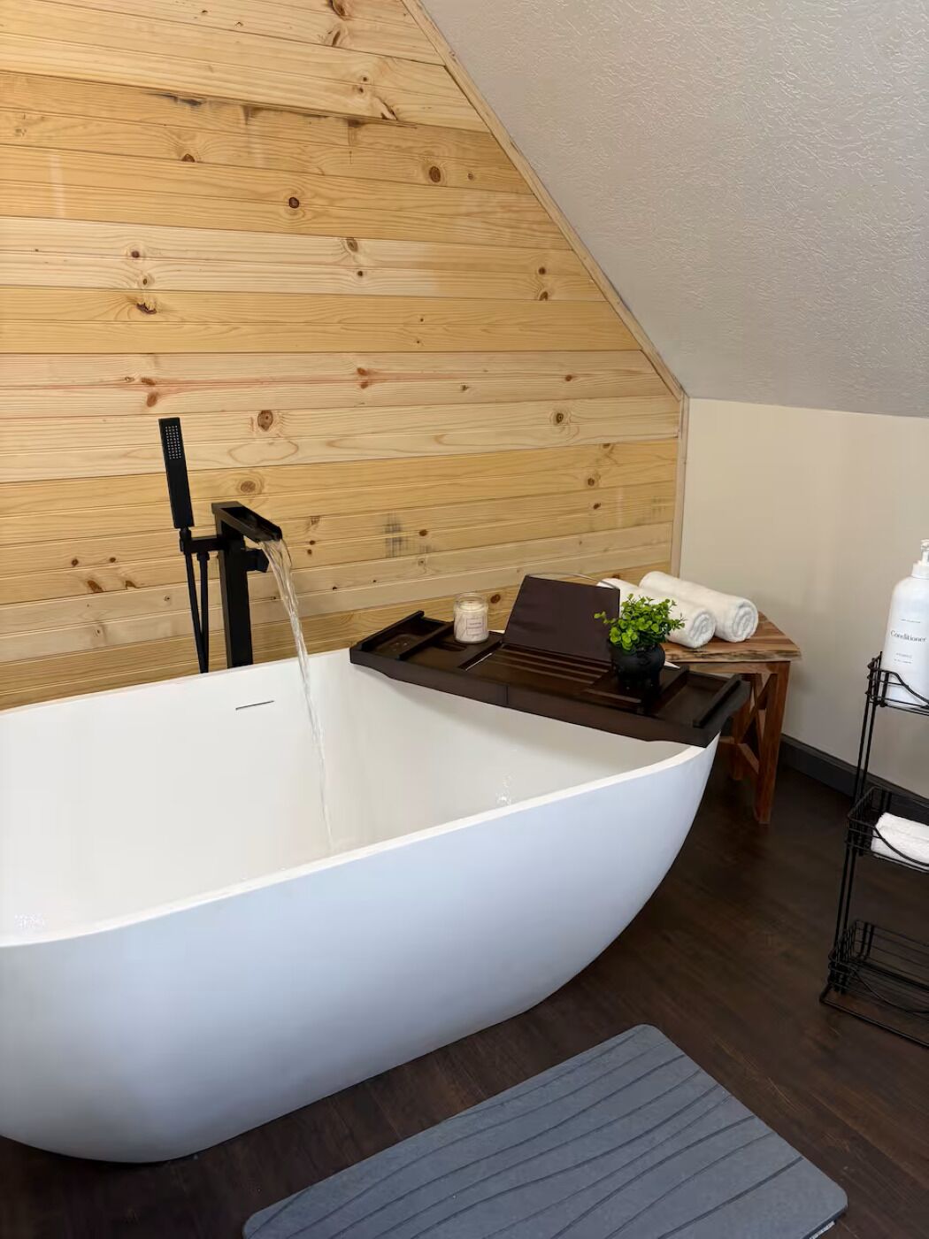 A modern bathroom with a freestanding bathtub, wooden wall paneling, a small table with a plant, and a shelf with toiletries.