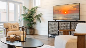 Ein Wohnzimmer mit Fernseher, der einen Sonnenuntergang zeigt, einer Couch mit Kissen, einer Zimmerpflanze und einer Anrichte.