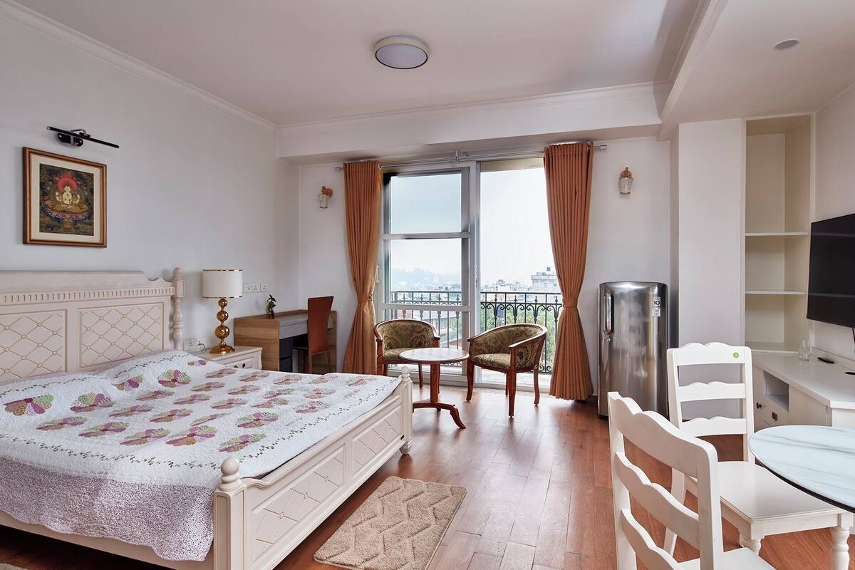 A hotel room with a bed, bedside table, lamp, chair, desk, TV, and a balcony with curtains.