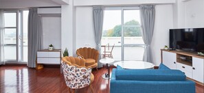 A modern living room with a sofa, a chair, a TV, a small table, and a view of the outdoors through large windows. - Luminous Apartments & Suites (Kathmandu)