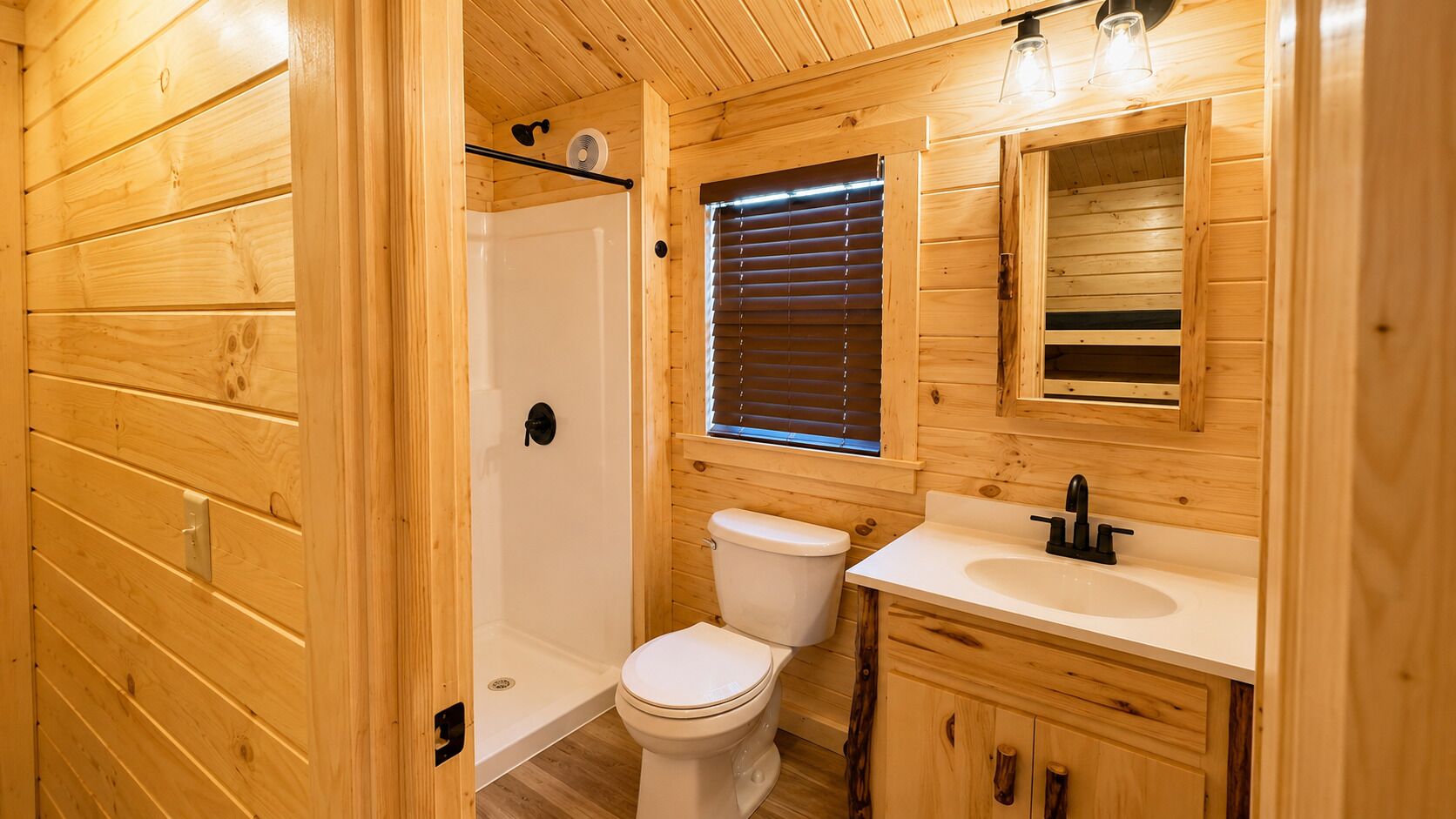 A wooden cabin bathroom with a shower, toilet, sink, and a small window with blinds.