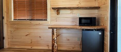 A compact kitchen with wooden walls, a built-in microwave, a small refrigerator, and a window with blinds.