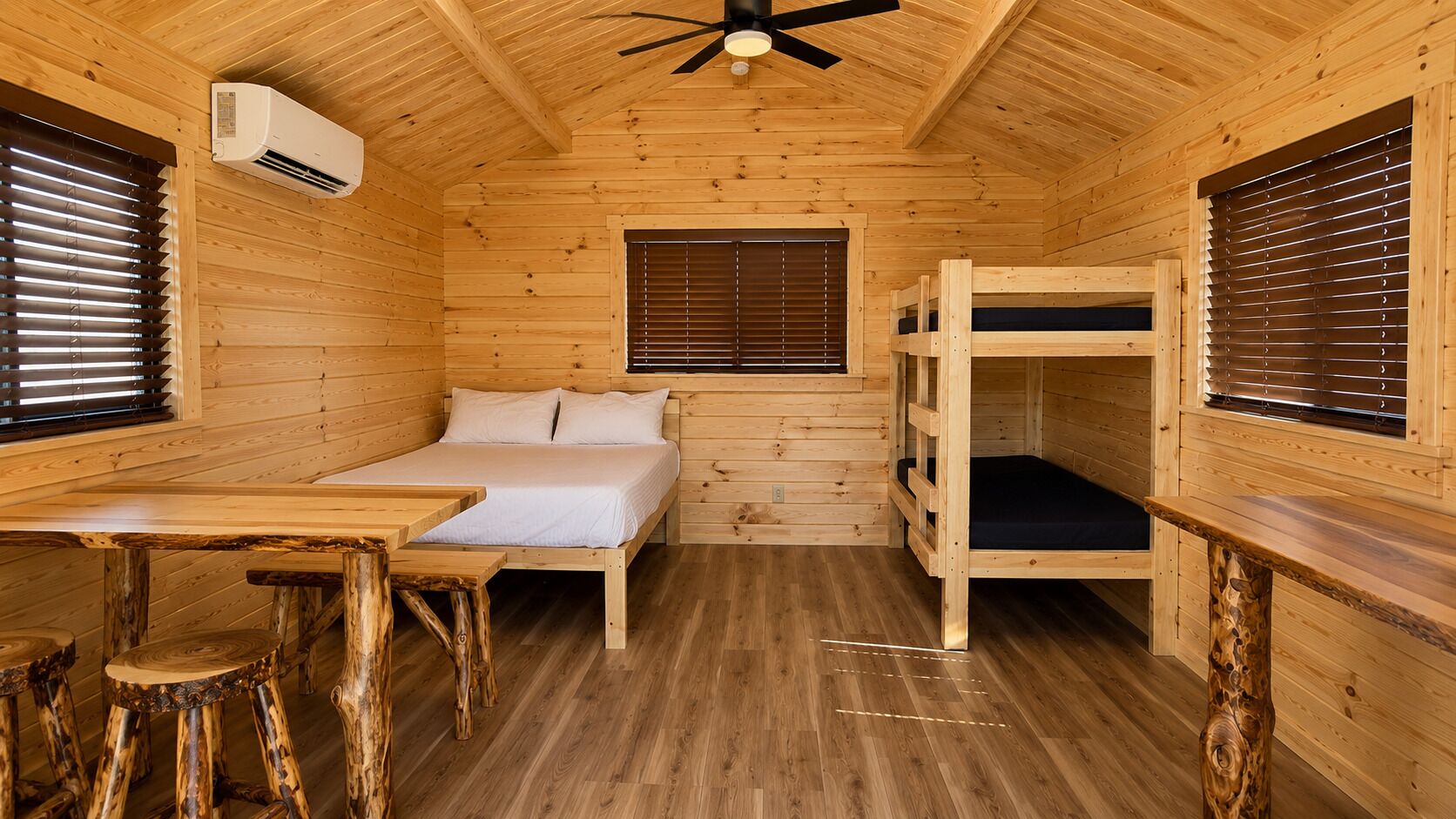 A wooden cabin interior with a bunk bed, a single bed, a table, and a ceiling fan.