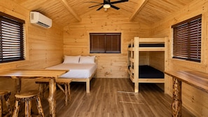L’intérieur d’une cabane en bois, avec un lit superposé, un lit simple, une table et un ventilateur de plafond.