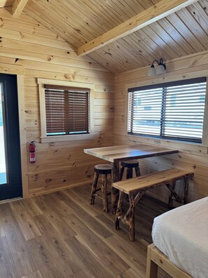 Interior de una cabaña de madera con una mesa de comedor, taburetes y dos ventanas con persianas.