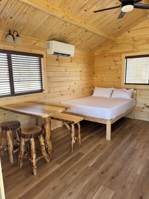 Interior de una cabaña de madera con una cama, una mesa, dos taburetes y un ventilador de techo.