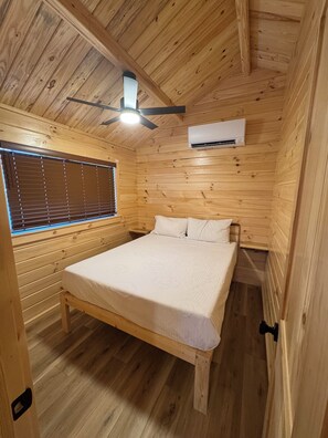 Un dormitorio de cabaña de madera con una cama, una ventana con persianas, un ventilador de techo y un equipo de aire acondicionado.