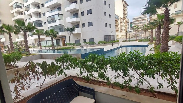 A balcony with two chairs and a table, overlooking a swimming pool and a multi-story building with balconies. - Quiet, Modern Studio Apartment in Zenata with Pool View (Âïn-Harrouda)