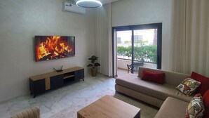 A modern living room with a flat-screen TV, a wooden coffee table, a sofa, and a sliding glass door leading to a balcony. - Quiet, Modern Studio Apartment in Zenata with Pool View (Âïn-Harrouda)