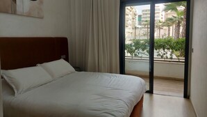 A bedroom with a bed, a window, and a view of buildings and greenery. - Quiet, Modern Studio Apartment in Zenata with Pool View (Âïn-Harrouda)