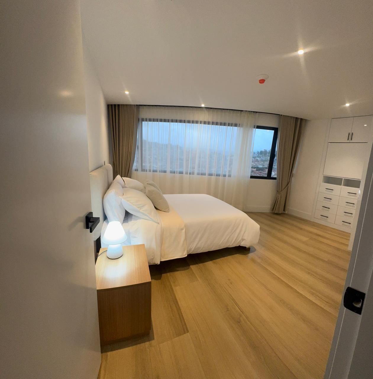 A bedroom with a bed, bedside table, lamp, window with curtains, and a wardrobe.