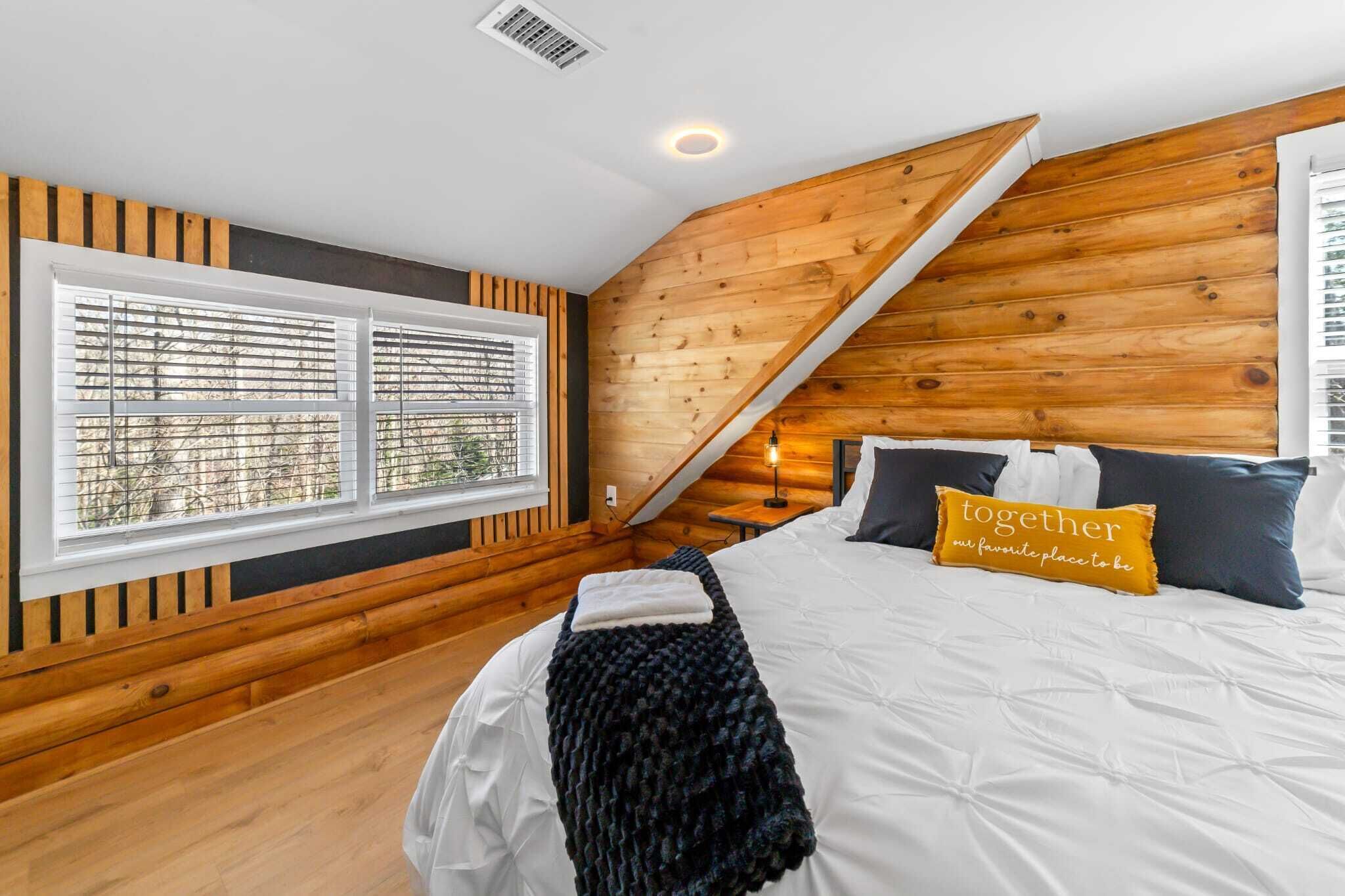 Une chambre avec un plafond en pente, des murs recouverts de lambris en bois, une grande fenêtre avec des stores, un lit avec une literie blanche et un oreiller orné d’un motif.
