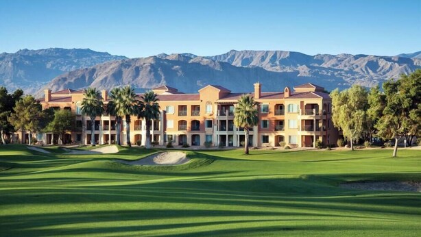 Un hotel de varios pisos con un campo de golf al frente, rodeado de montañas.