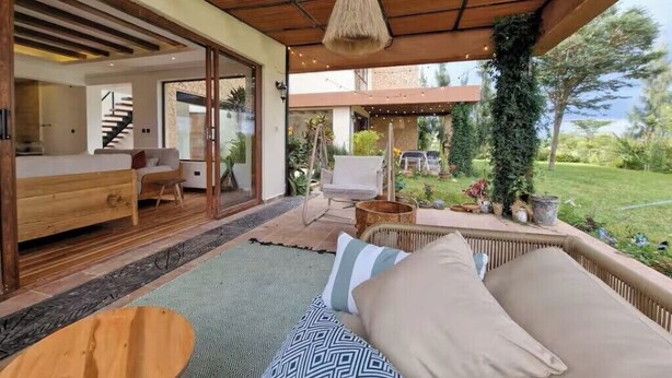 Un'area relax esterna moderna con tetto in legno, un divano con cuscini decorati e vista su un giardino e un edificio sullo sfondo.