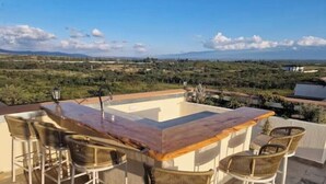 Zona bar esterna con piscina, circondata da poltrone in vimini e vista su un paesaggio con colline lontane.