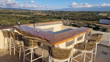 An outdoor bar area with a pool, surrounded by wicker chairs, overlooking a landscape with distant hills.