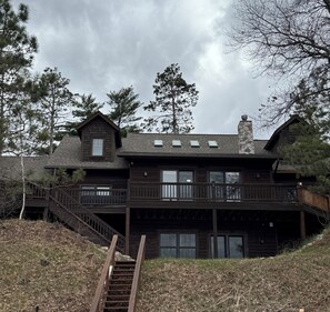A two-story log cabin with a stone chimney, a wooden deck, and a staircase leading to the entrance.