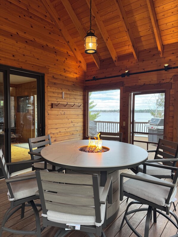 A wooden interior with a round table, chairs, a fire pit, and a view of a body of water.
