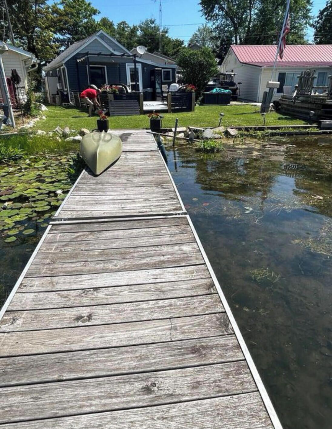 En træbro fører til et hus med parabol, omgivet af grøn vegetation og en lille sø.