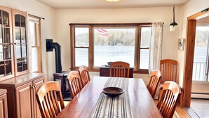 Une salle à manger avec une grande table en bois, des chaises, un poêle noir et une vue sur un plan d’eau, visible à travers de grandes fenêtres.