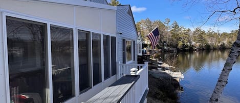 A waterfront house with a deck, large windows, and an American flag.