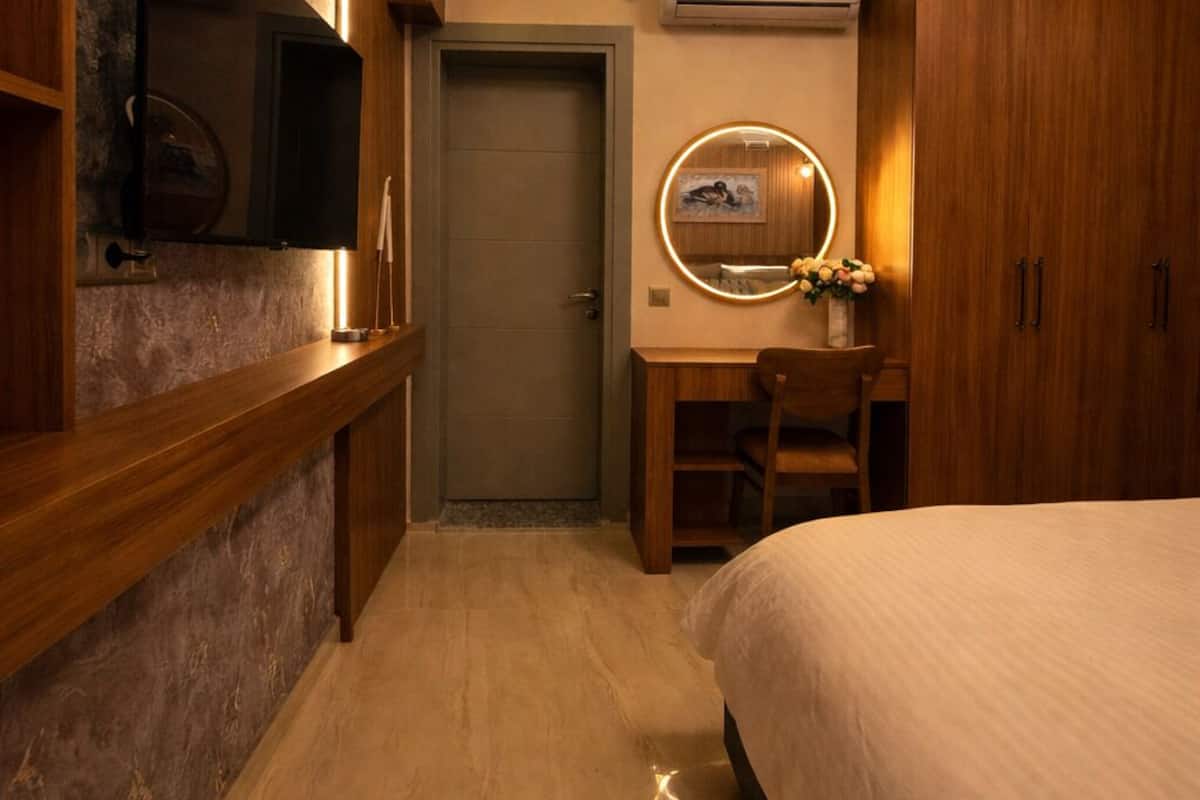 Une chambre à coucher comprenant un lit, un bureau avec une chaise, un téléviseur fixé au mur, un miroir rond et un appareil de climatisation.