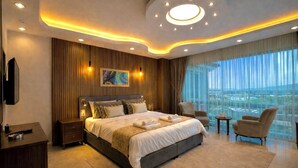 A modern hotel room with a large bed, a TV, a desk, and a view of the city. - Villa Nuri Konaklama (Antakya)