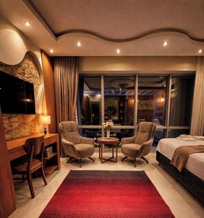Quarto de hotel com televisão, duas poltronas, uma mesa pequena com um vaso, uma cama e uma janela grande.