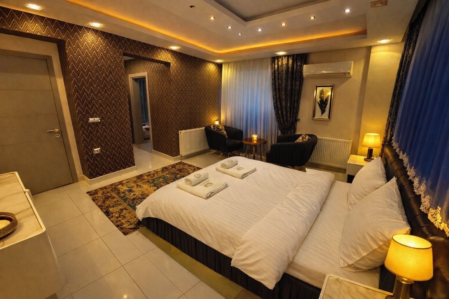 A hotel room with a large bed, a desk, a chair, a small table, and a window with curtains.