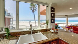 A kitchen with a large window overlooking a beach, a sink, and a dining area with chairs. - *New* Ocean Surf View | 2nd Floor Condo w/ Parking (San Diego)