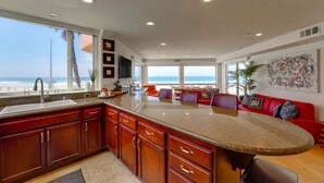 A kitchen with a large island, a dining area with a red sofa, and a view of the beach. - *New* Ocean Surf View | 2nd Floor Condo w/ Parking (San Diego)