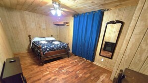 Ein Schlafzimmer mit Holzboden, einem Bett mit blumigem Überwurf, einem Deckenventilator, einem blauen Vorhang und einem Spiegel an der Wand.