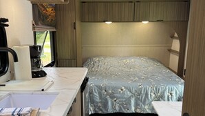 Interior compacto de uma motorhome com cozinha compacta, uma cama com roupa de cama floral e uma janela com vista para vegetação.