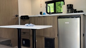 A compact kitchenette with a refrigerator, a coffee maker, and a window. - New Northwoods Escape!  2026 Wildwood FSX Camper Near Bike Trails, Lakes & Pines (Boulder Junction)